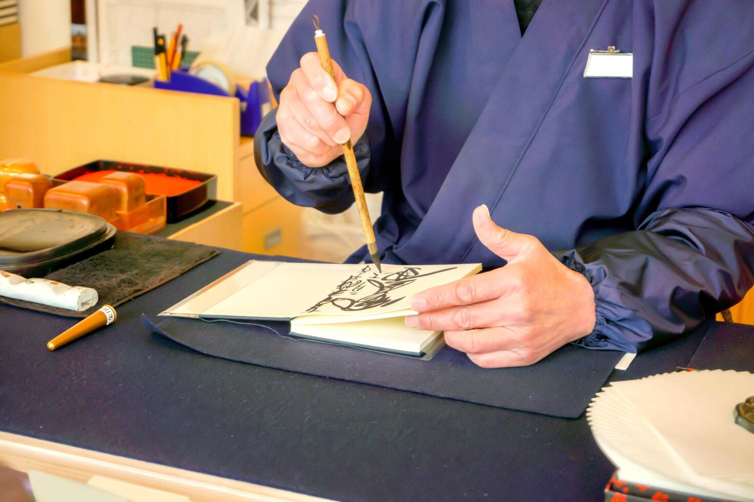 Nahansicht Hand von Person tragen ein Blau National Kleid und halten ein Chinesisch Bürste Schreiben japanisch Texte von schwarz Tinte auf ein Notizbuch Papier. foto