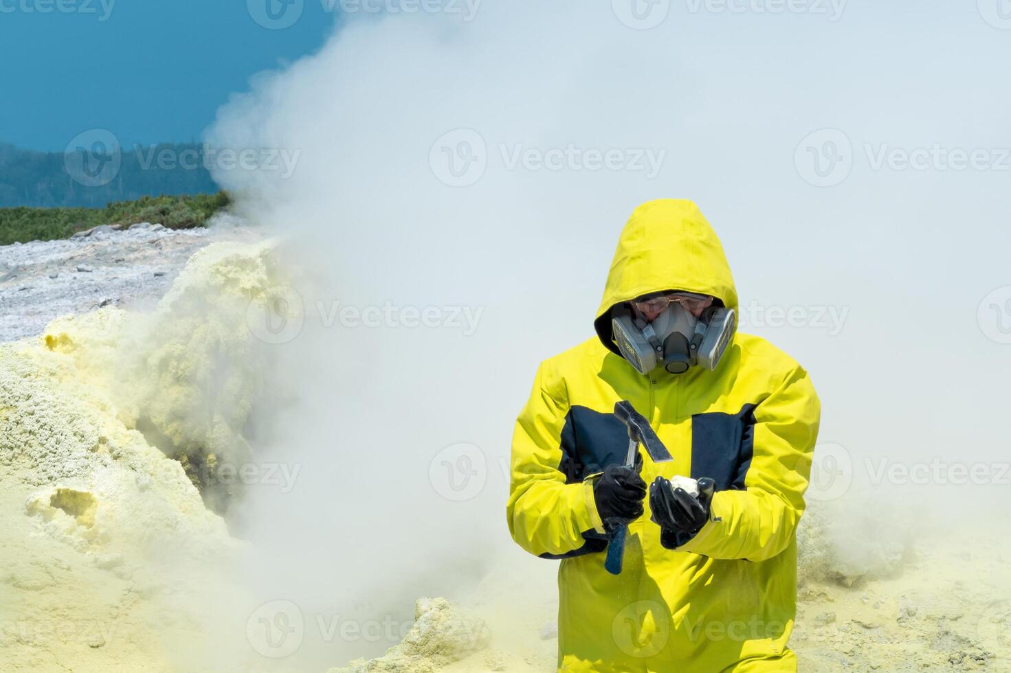 Mann Vulkanologe auf das Hintergrund von ein Rauchen Fumarole untersucht ein Stichprobe von ein Schwefel Mineral mit ein geologisch Hammer foto