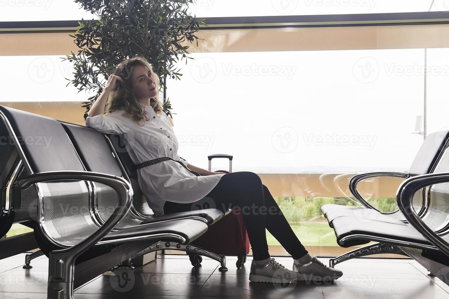 Frau Reisender mit Gepäck sitzt im das warten Zimmer von das Flughafen oder Zug Bahnhof und ist warten zum das Flug foto