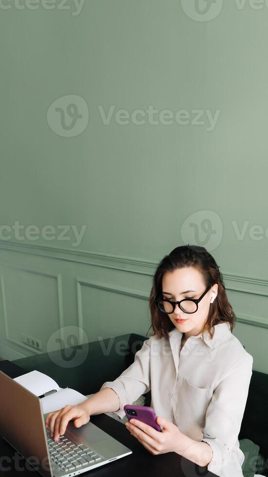 produktiv Gespräche. Frau balancieren Laptop und Smartphone im Video Forderung. Multitasking Freude. beschäftigt Frau im Brille jongliert Video Anruf auf Laptop und Smartphone mit ein Lächeln foto