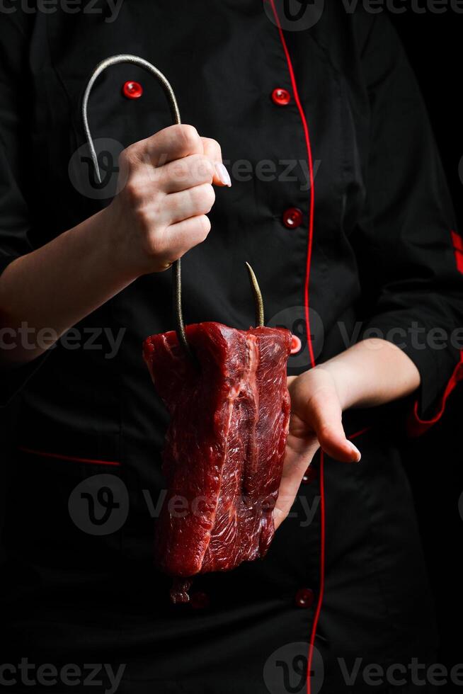 ein Metzger hält ein Stück von Kalbfleisch auf ein Metall Haken. Fleisch Vorbereitung. auf ein schwarz Hintergrund. foto