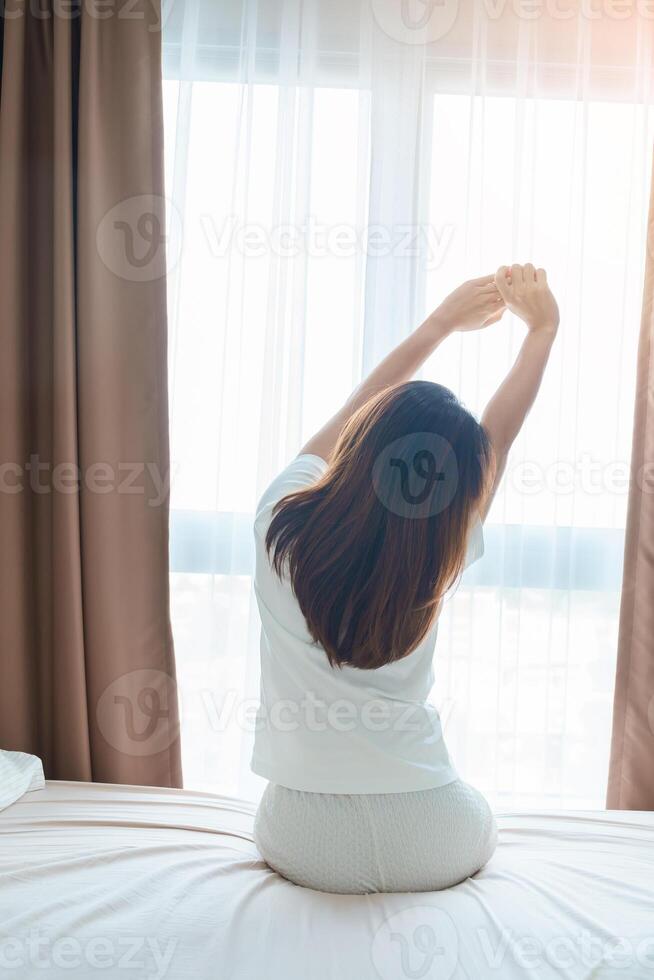 glücklich Frau Dehnen auf Bett nach aufwachen hoch, jung Erwachsene weiblich steigend Waffen und suchen zu Fenster im das früh Morgen. frisch entspannen und haben ein nett Tag Konzepte foto