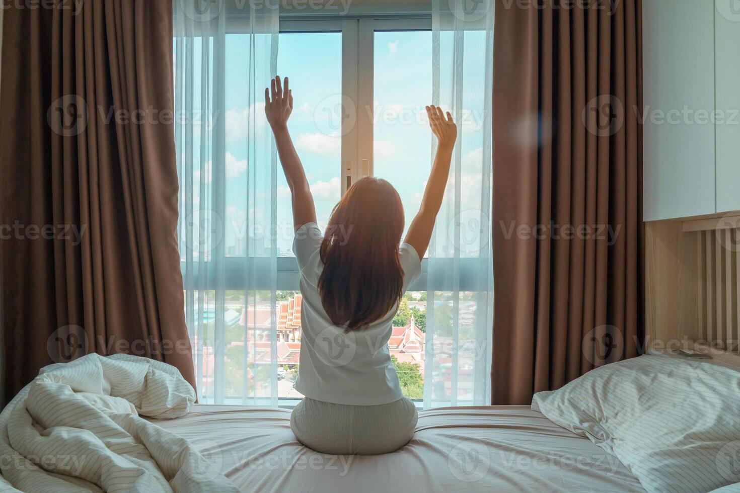 glücklich Frau Dehnen auf Bett nach aufwachen hoch, jung Erwachsene weiblich steigend Waffen und suchen zu Fenster im das früh Morgen. frisch entspannen und haben ein nett Tag Konzepte foto