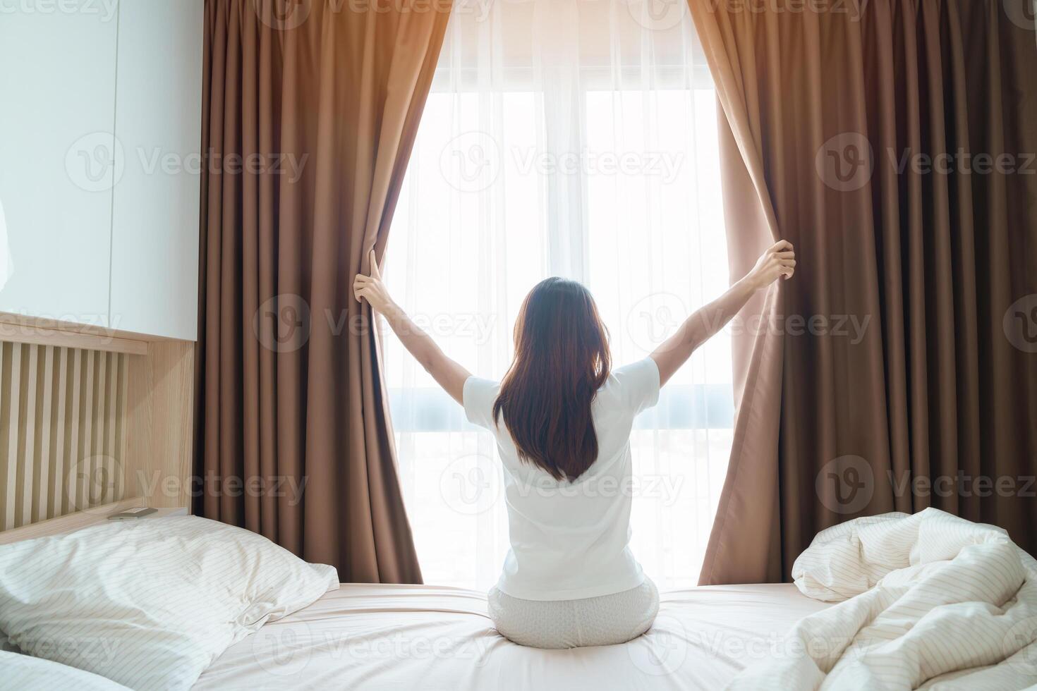 glücklich Frau Dehnen auf Bett nach aufwachen hoch, jung Erwachsene weiblich steigend Waffen und suchen zu Fenster im das früh Morgen. frisch entspannen und haben ein nett Tag Konzepte foto