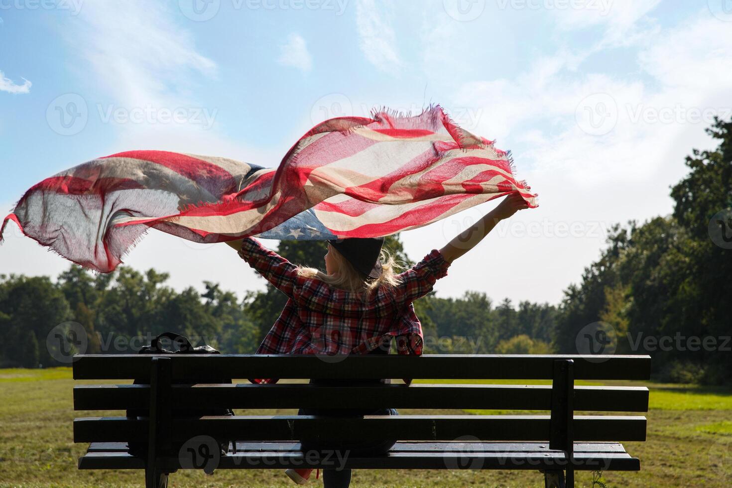 jung Mädchen sitzt auf Bank und hält ein amerikanisch Flagge winken im das Wind. hoch Qualität Foto