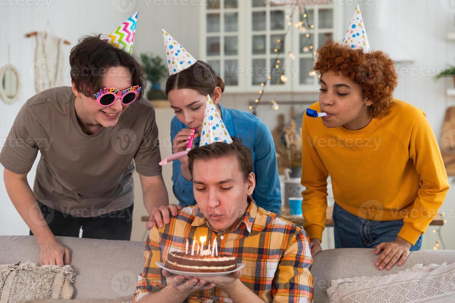 machen ein Wunsch. Mann tragen Party Deckel weht aus Verbrennung Kerzen auf Geburtstag Kuchen. glücklich Geburtstag Party. Gruppe von freunde wünscht sich Kerl glücklich Geburtstag. Menschen feiern Geburtstag mit Party beim Zuhause foto