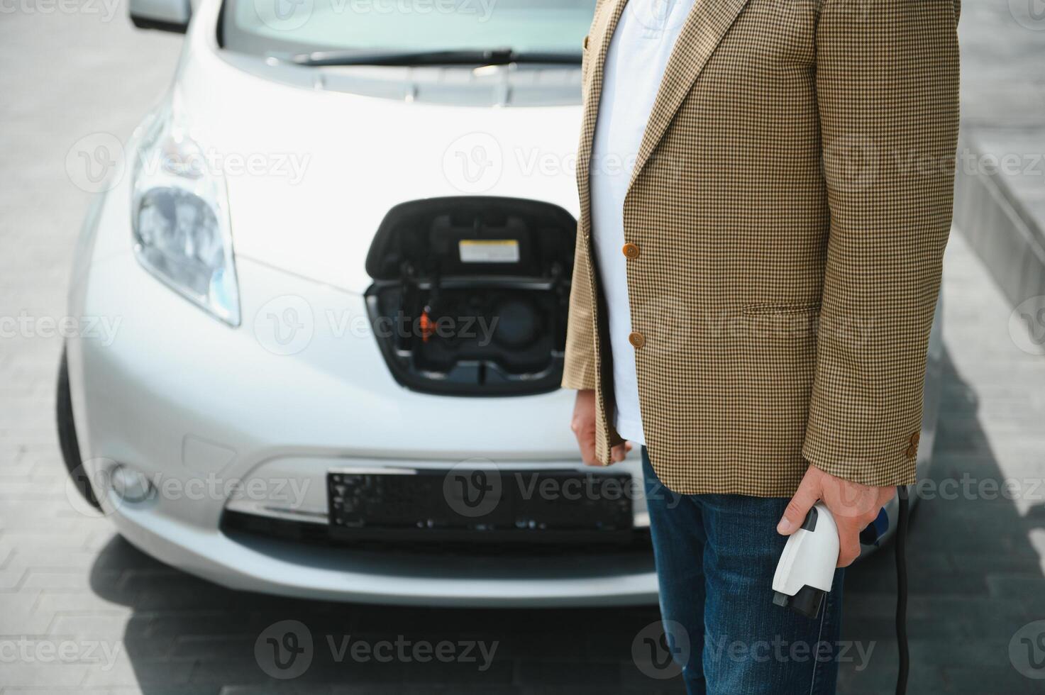 gut aussehend Mann halten Laden Kabel beim elektrisch Laden Bahnhof Punkt Stehen in der Nähe von seine Neu Wagen. foto