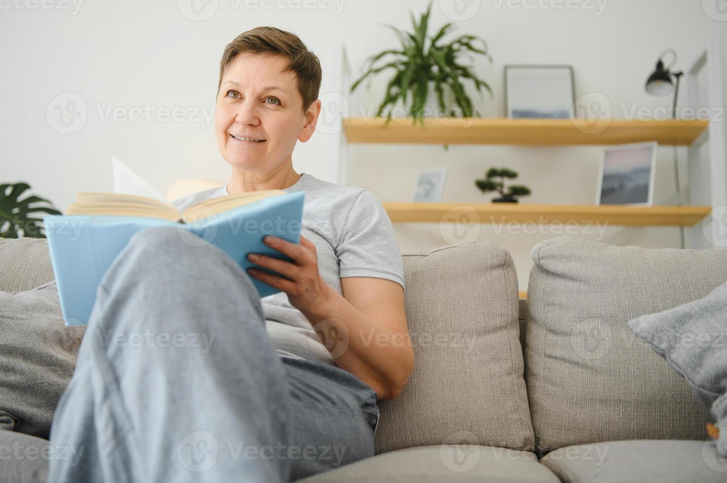Porträt von ein glücklich reifen Frau entspannend auf Couch und lesen ein Buch im ein modern Haus foto