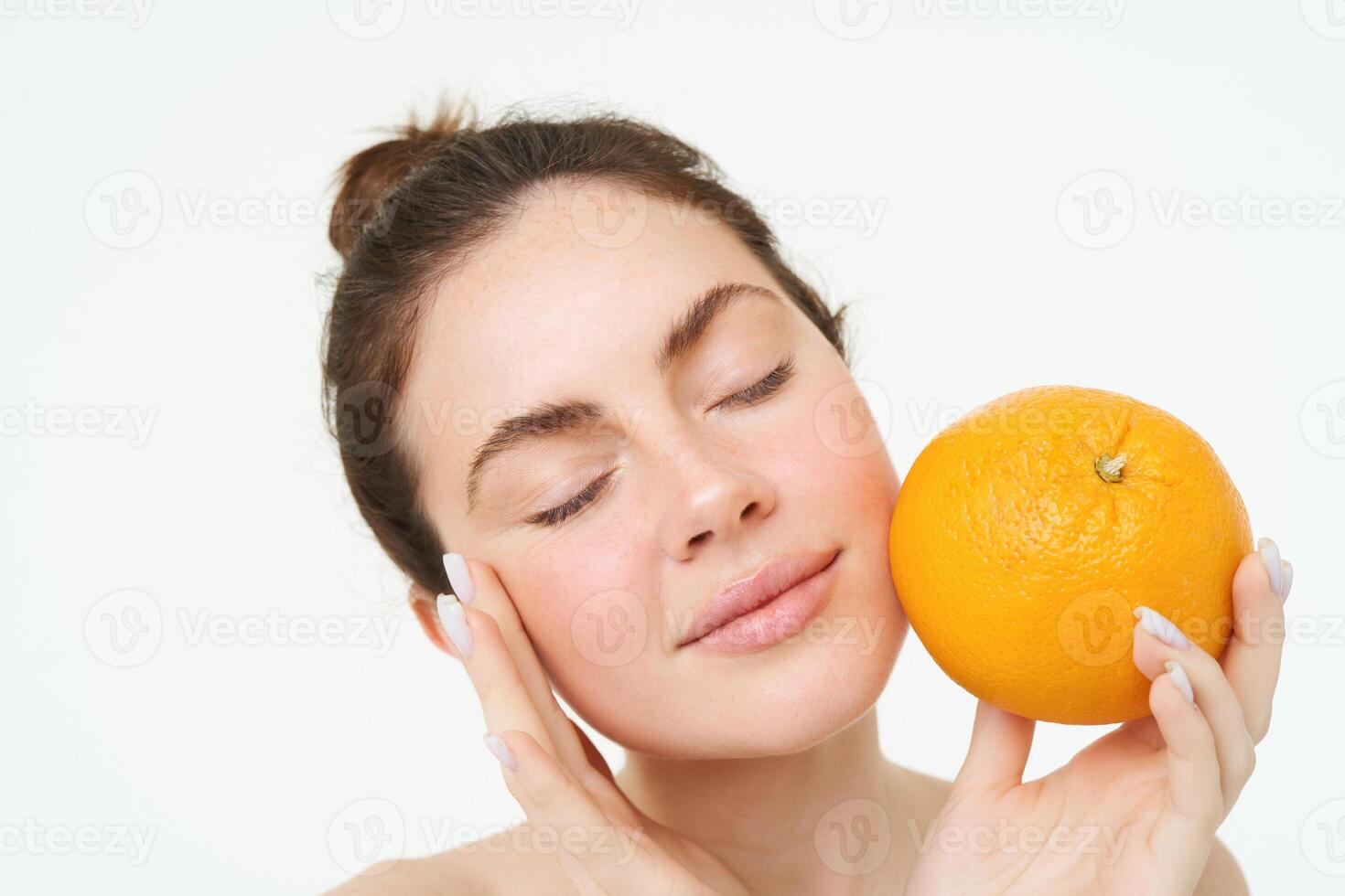 Porträt von jung Frau halten Orange in der Nähe von Gesicht ohne Schönheitsfehler, lächelnd, Konzept von Vitamine, Nahrung und organisch Gesichts- Behandlungen, Weiß Hintergrund foto