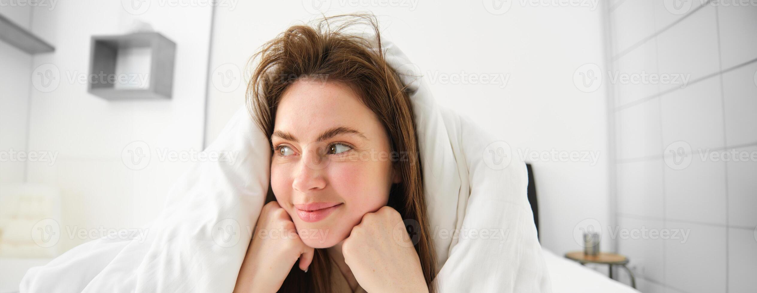 süß Mädchen mit unordentlich Haar, Lügen im Bett bedeckt im Weiß Blätter Bettdecke, lächelnd und Lachen kokett, Ausgaben Zeit im ihr Schlafzimmer foto