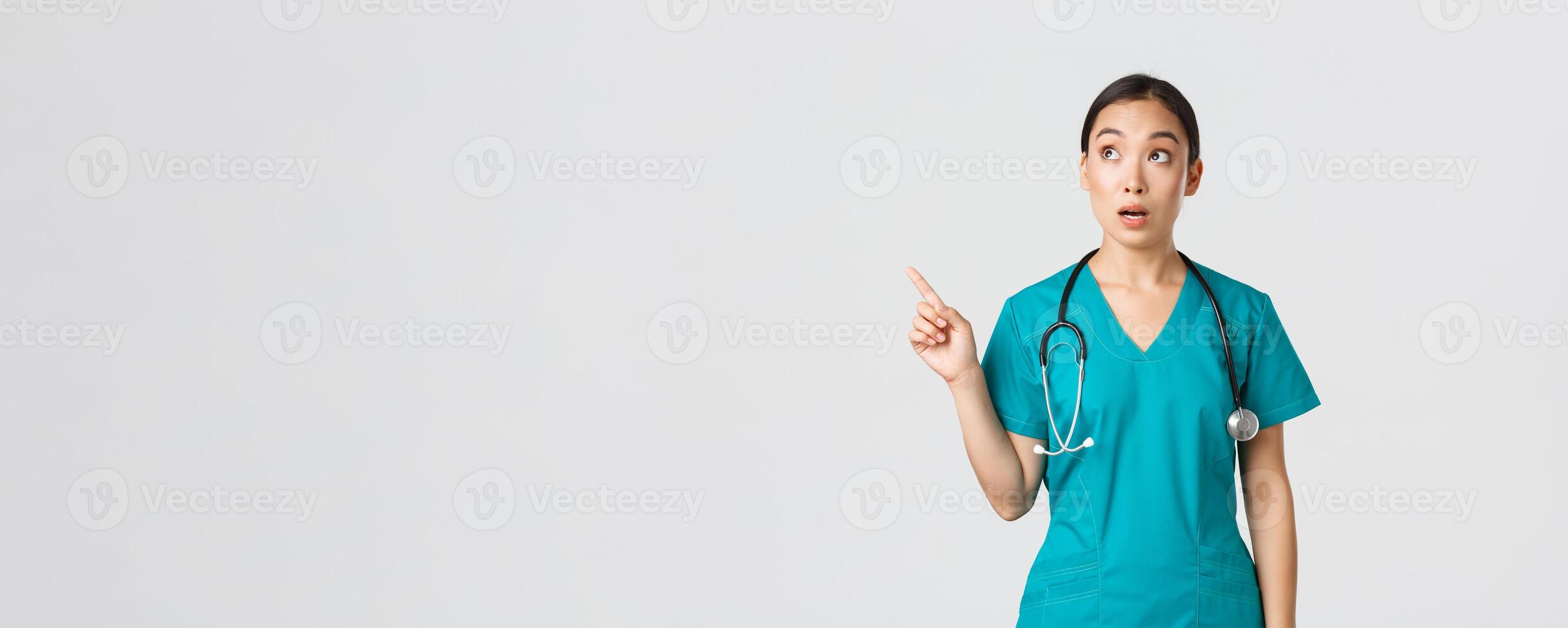 COVID-19, Gesundheitswesen Arbeitskräfte, Pandemie Konzept. überrascht und erstaunt weiblich asiatisch Arzt suchen und zeigen Oberer, höher links Ecke. erstaunt Koreanisch Arzt starren beim Banner mit Werbung foto