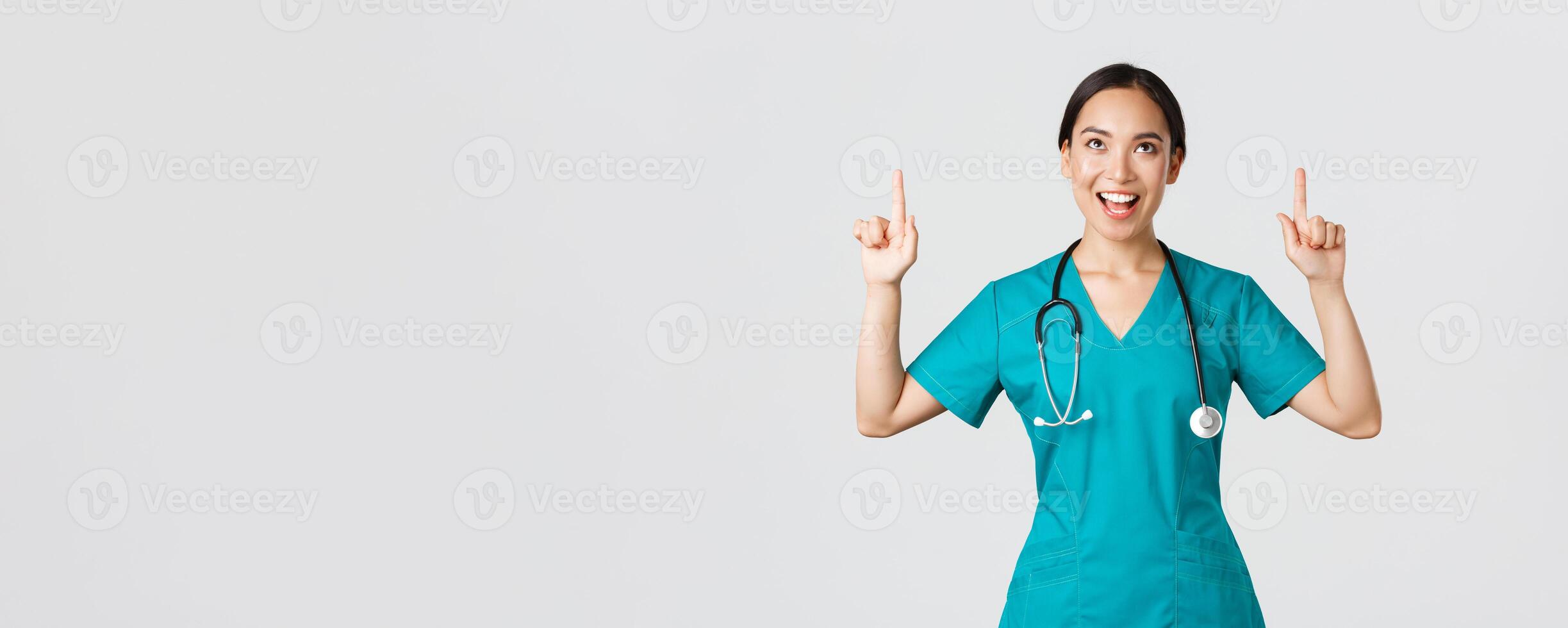 COVID-19, Gesundheitswesen Arbeitskräfte, Pandemie Konzept. amüsiert heiter asiatisch weiblich Krankenschwester, Arzt oder der Chirurg im Peelings zeigen Finger und suchen oben mit zufrieden Gesicht, Stehen Weiß Hintergrund foto