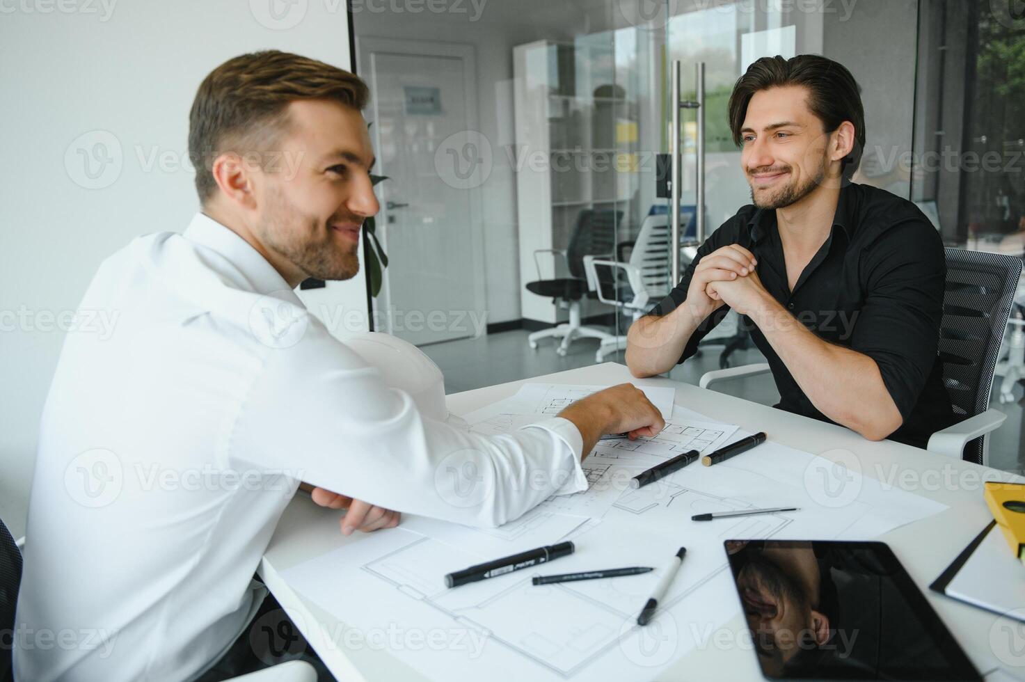 zwei Gebäude Designer Stehen im ein modern Büro gelehnt Über ein Schreibtisch diskutieren Blaupausen zusammen foto