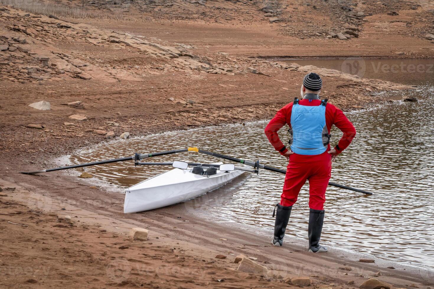 Senior männlich Ruderer tragen Trockenanzug mit ein Küsten Rudern Schale auf ein Ufer von Pferdezahn Reservoir im fallen oder Winter Landschaft. foto