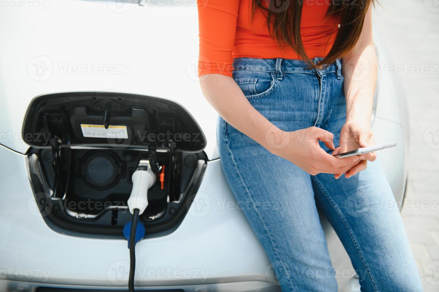 mit Smartphone während warten. Frau auf das elektrisch Autos aufladen Bahnhof beim Tageszeit. Marke Neu Fahrzeug. foto