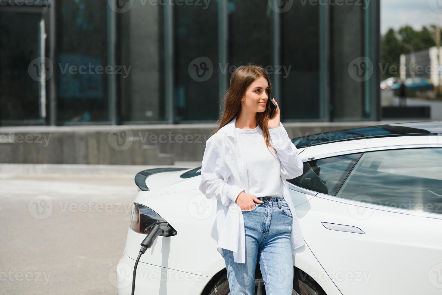 Telefon im Hände. Frau auf das elektrisch Autos aufladen Bahnhof beim Tageszeit. Marke Neu Fahrzeug. foto