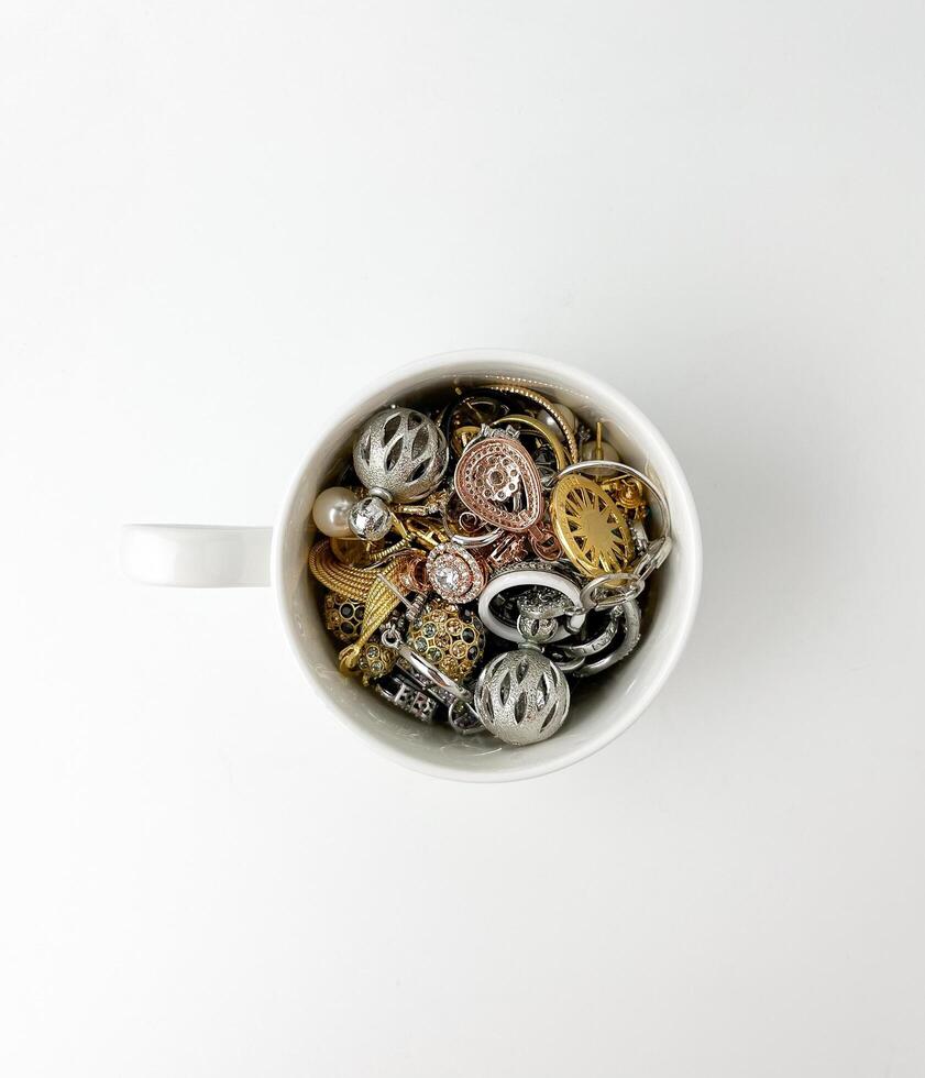 ein Bündel von Metall Gold Silber Material Ohrringe, Halskette, und Ringe im Weiß Keramik Glas Tasse. Erbe Schmuck Mode Objekt Fotografie isoliert auf oben eben legen Weiß Studio Hintergrund. foto