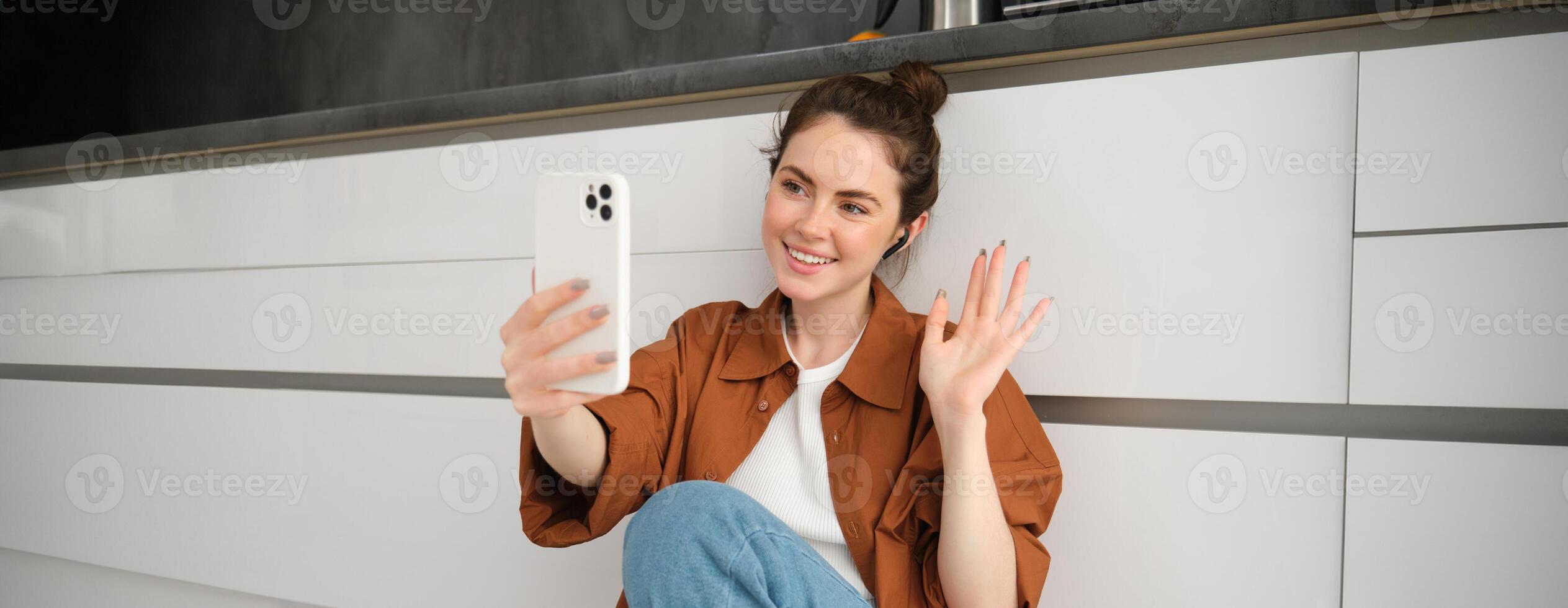 Porträt von glücklich lächelnd Frau Video chatten, sitzt auf Küche Fußboden beim heim, reden zu Freund auf Sozial Medien App mit Smartphone foto