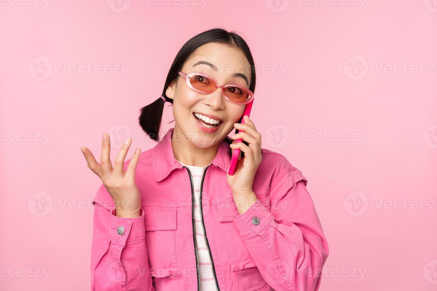 schön Koreanisch weiblich Modell- im Sonnenbrille, reden auf Handy, Mobiltelefon Telefon mit glücklich Gesicht, mit zellular Bedienung zu Anruf Freund auf Smartphone, Stehen Über Rosa Hintergrund foto