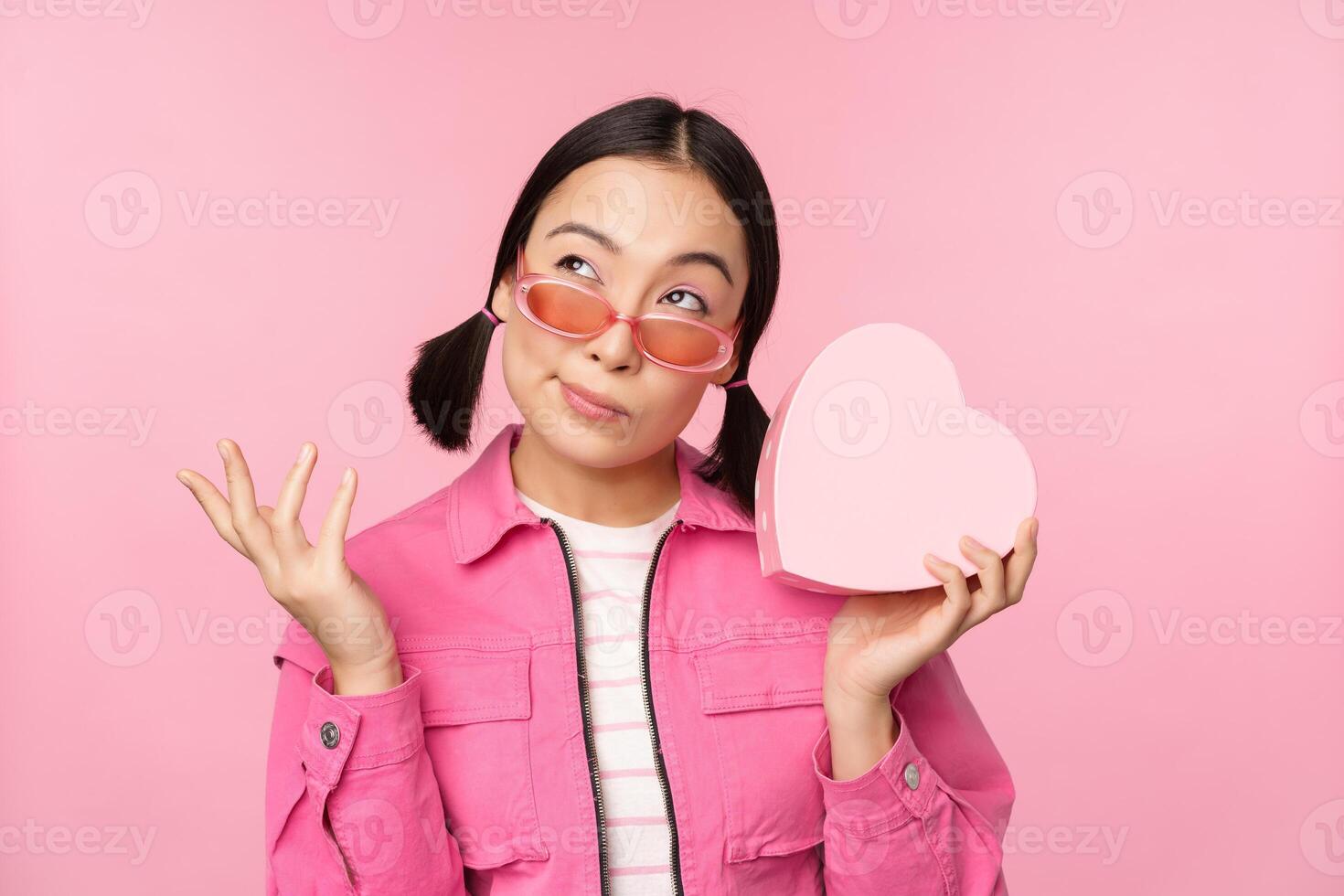 Bild von stilvoll asiatisch Freundin im Sonnenbrille, raten was ist Innerhalb Geschenk Kasten, herzförmig gegenwärtig, Stehen Über Rosa Hintergrund foto