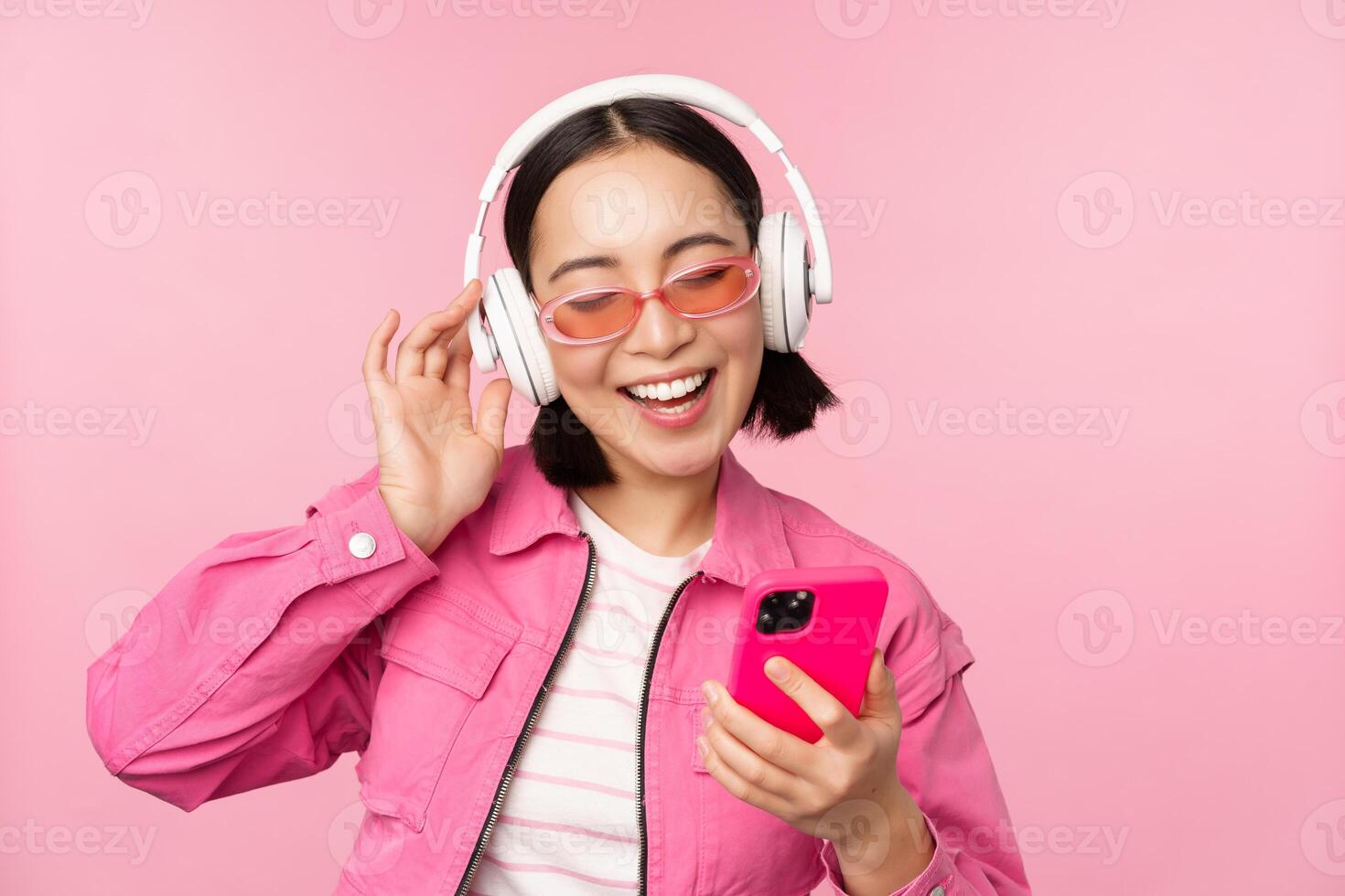 stilvoll asiatisch Mädchen Tanzen mit Smartphone, Hören Musik- im Kopfhörer auf Handy, Mobiltelefon Telefon Anwendung, lächelnd und Lachen, posieren gegen Rosa Hintergrund foto