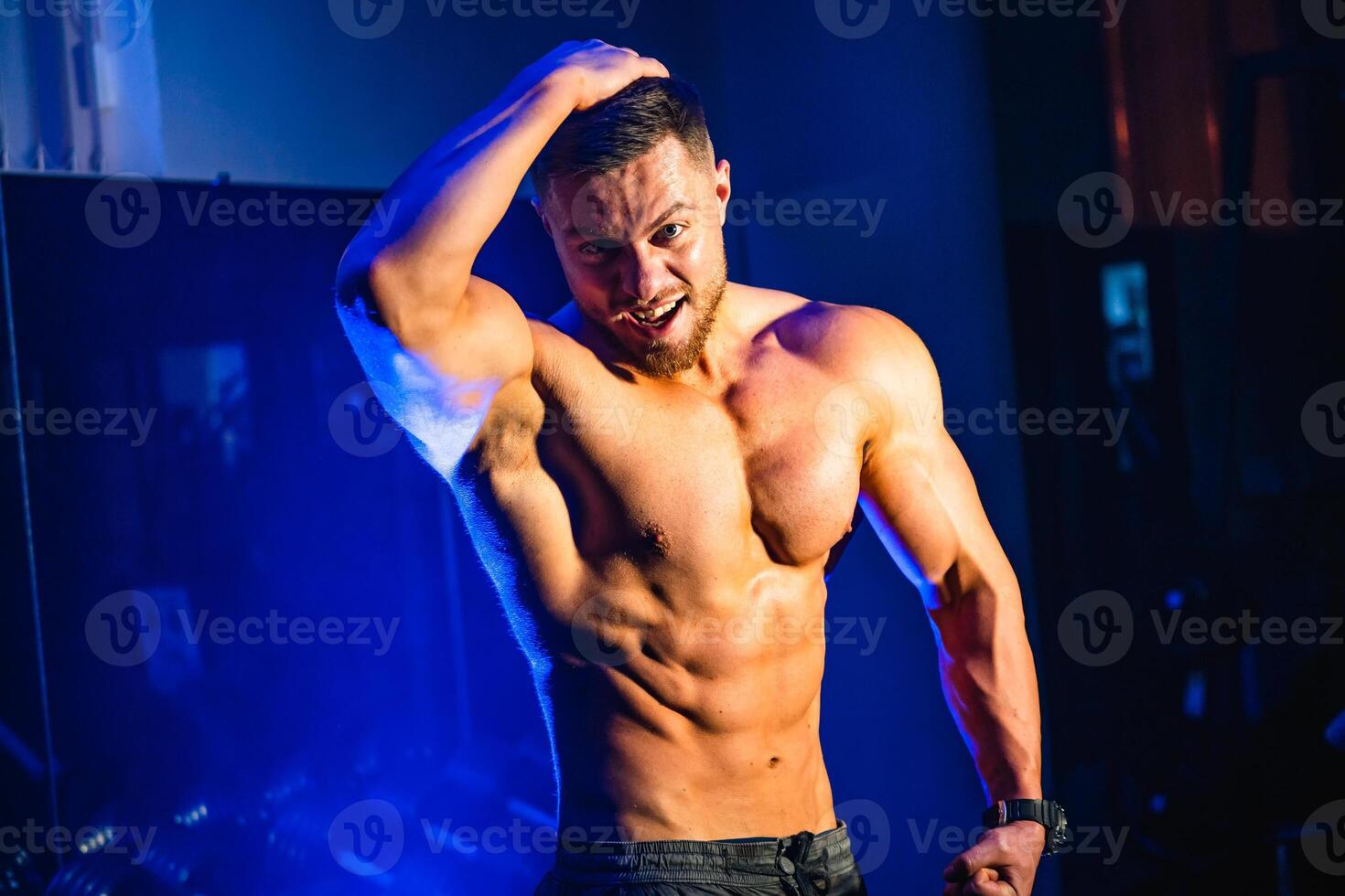 gut aussehend Mann mit groß Muskeln, posieren beim das Kamera im das Fitnessstudio, schwarz und Blau Hintergrund. Porträt von ein lächelnd Bodybuilder. Nahaufnahme. foto