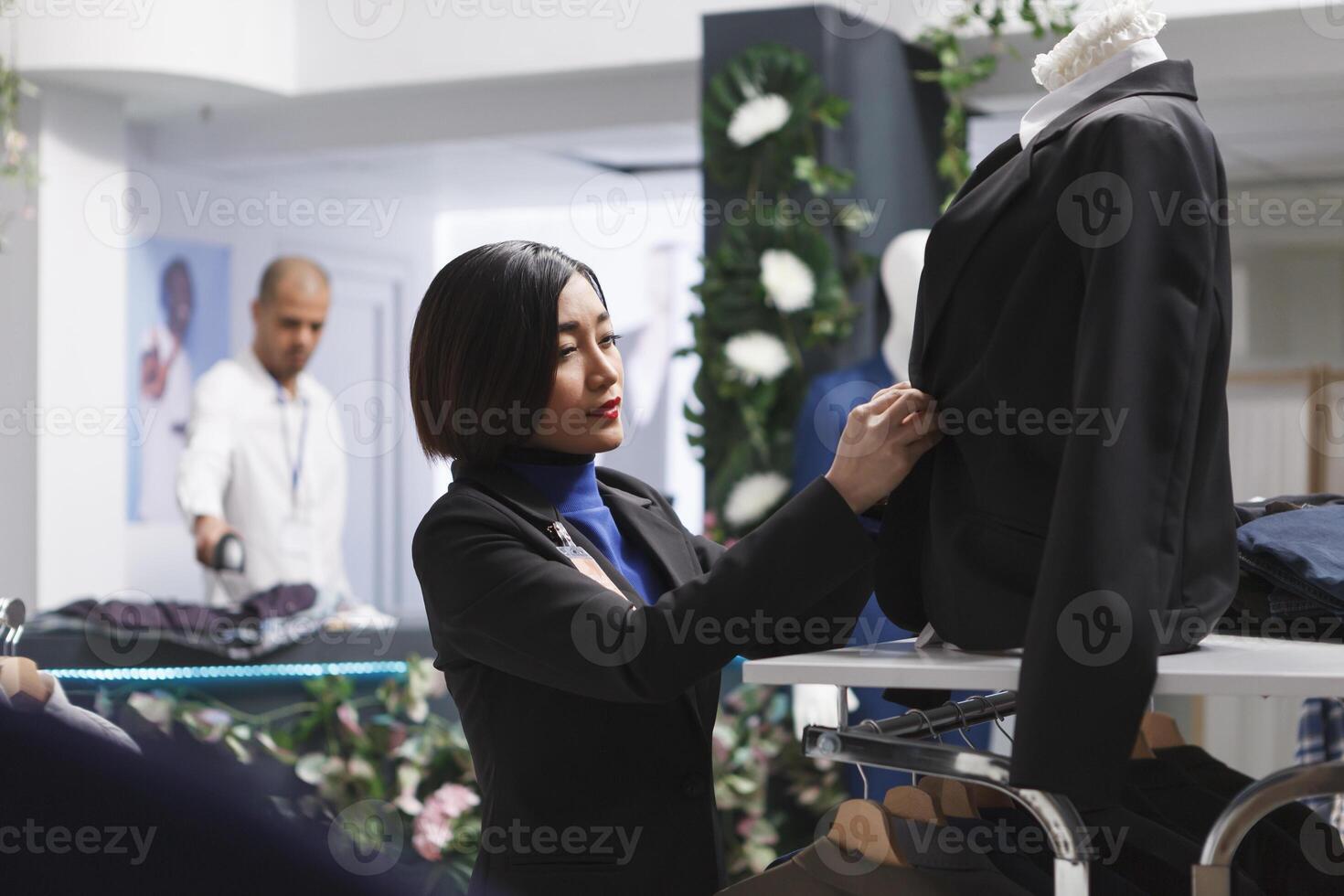 Kleidung Geschäft asiatisch Frau Assistent Prüfung Jacke auf Mannequin während Putten auf passen auf Mannequin. Boutique Verkäufer Erstellen stilvoll formal Outfit zu Vitrine bekleidung Marke foto