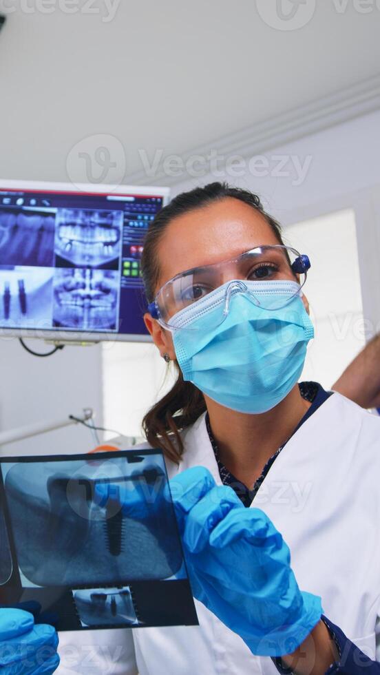 geduldig pov im Dental Büro Planung Chirurgie Schritte von Zähne Hohlraum, Zahnarzt zeigen auf Röntgen Bild. Stomatologie Arzt tragen schützend Maske und Handschuhe, Arbeiten im modern stomatologisch Klinik foto