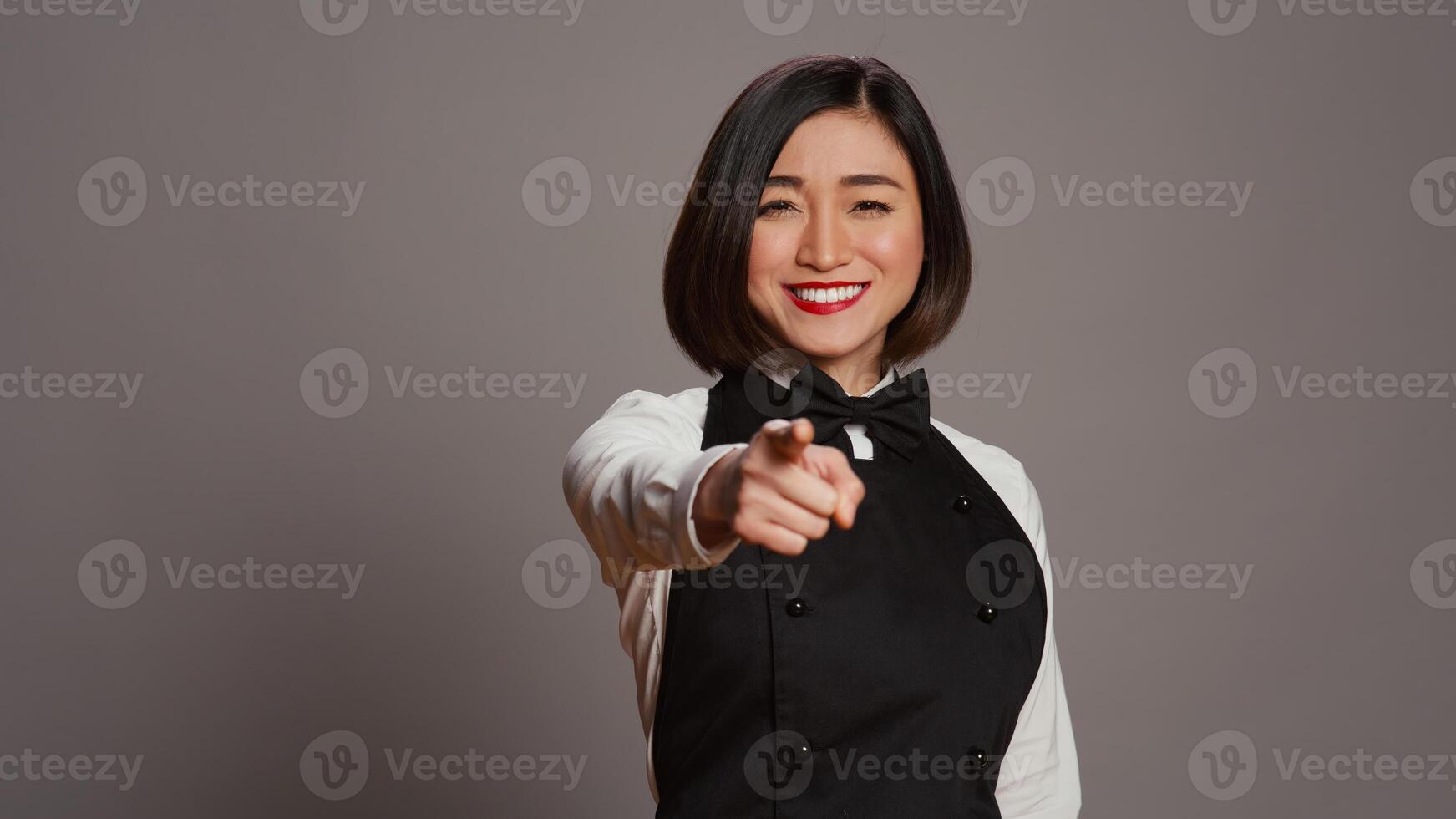 asiatisch Kellnerin zeigen beim Kamera zu wählen Sie Über grau Hintergrund, tragen Uniform mit Bogen und Schürze. Frau Gastronomie Arbeiter wählen jemand zu Arbeit beim ein Restaurant. Kamera b. foto