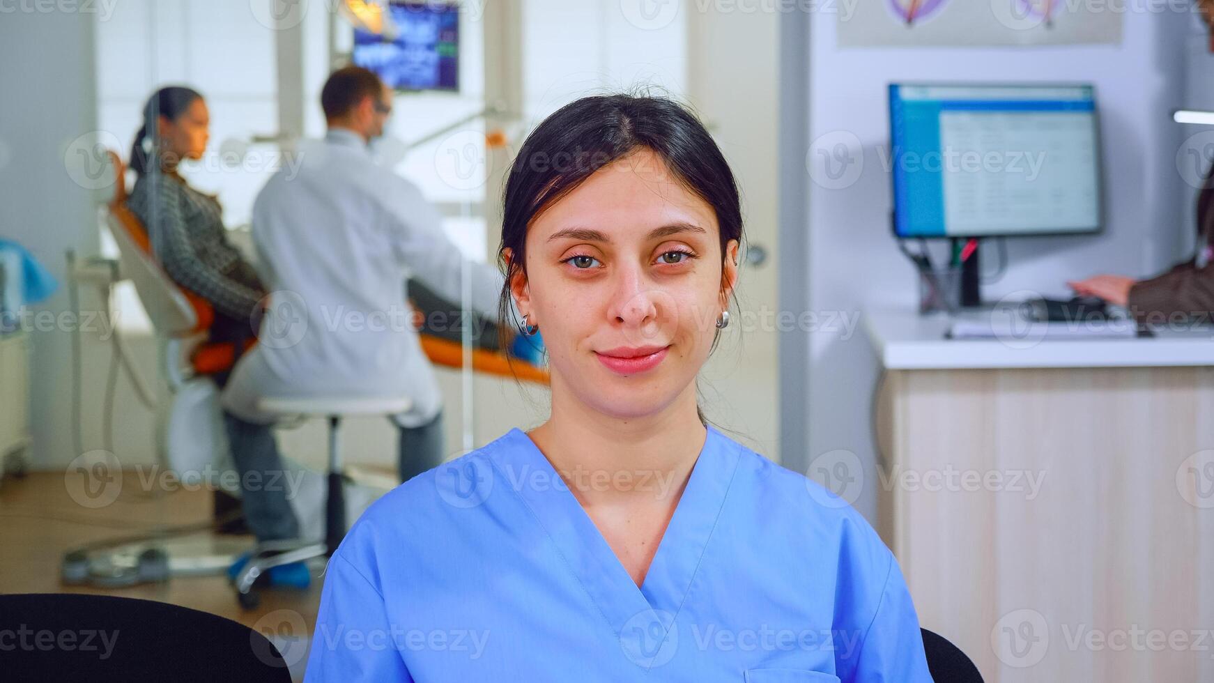 Dental Assistent suchen beim Kamera während Arzt Prüfung geduldig im Hintergrund. Fachmann Stomatologe Krankenschwester lächelnd auf Webcam Sitzung auf Stuhl im warten Zimmer von stomatologisch Klinik. foto