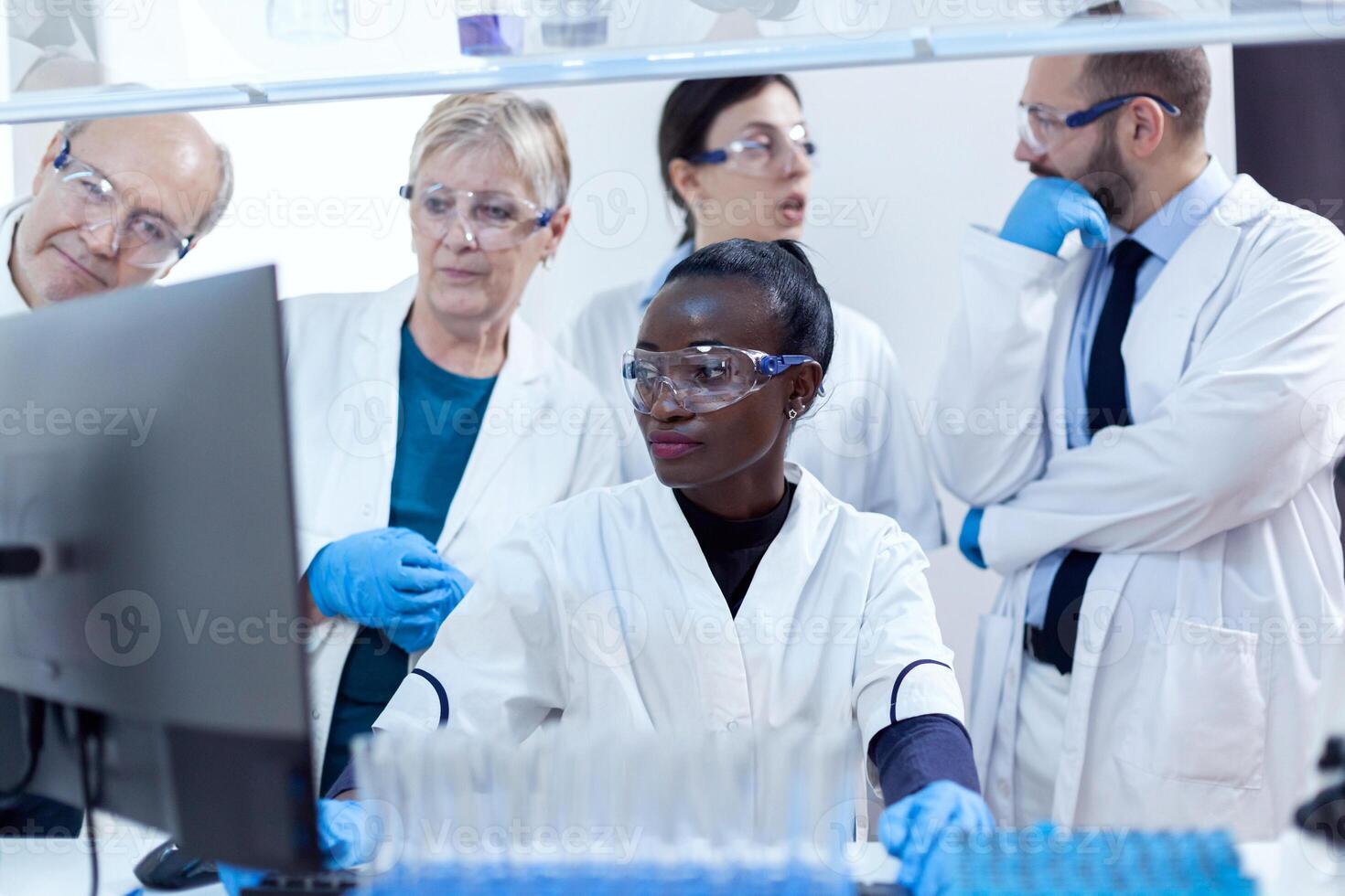 Mannschaft von Wissenschaftler mit Multiethnizität diskutieren chemisch Formel im Vorderseite von Computer. schwarz Gesundheitswesen Forscher im Biochemie Labor tragen steril Ausrüstung. foto