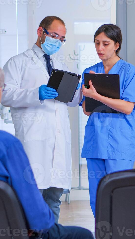 Dental Spezialist Sprechen mit Assistent Stehen im warten Zimmer von stomatologisch Klinik Vor Patienten Untersuchung, Krankenschwester nehmen Anmerkungen auf Zwischenablage, während Arzt suchen auf Tablette. foto