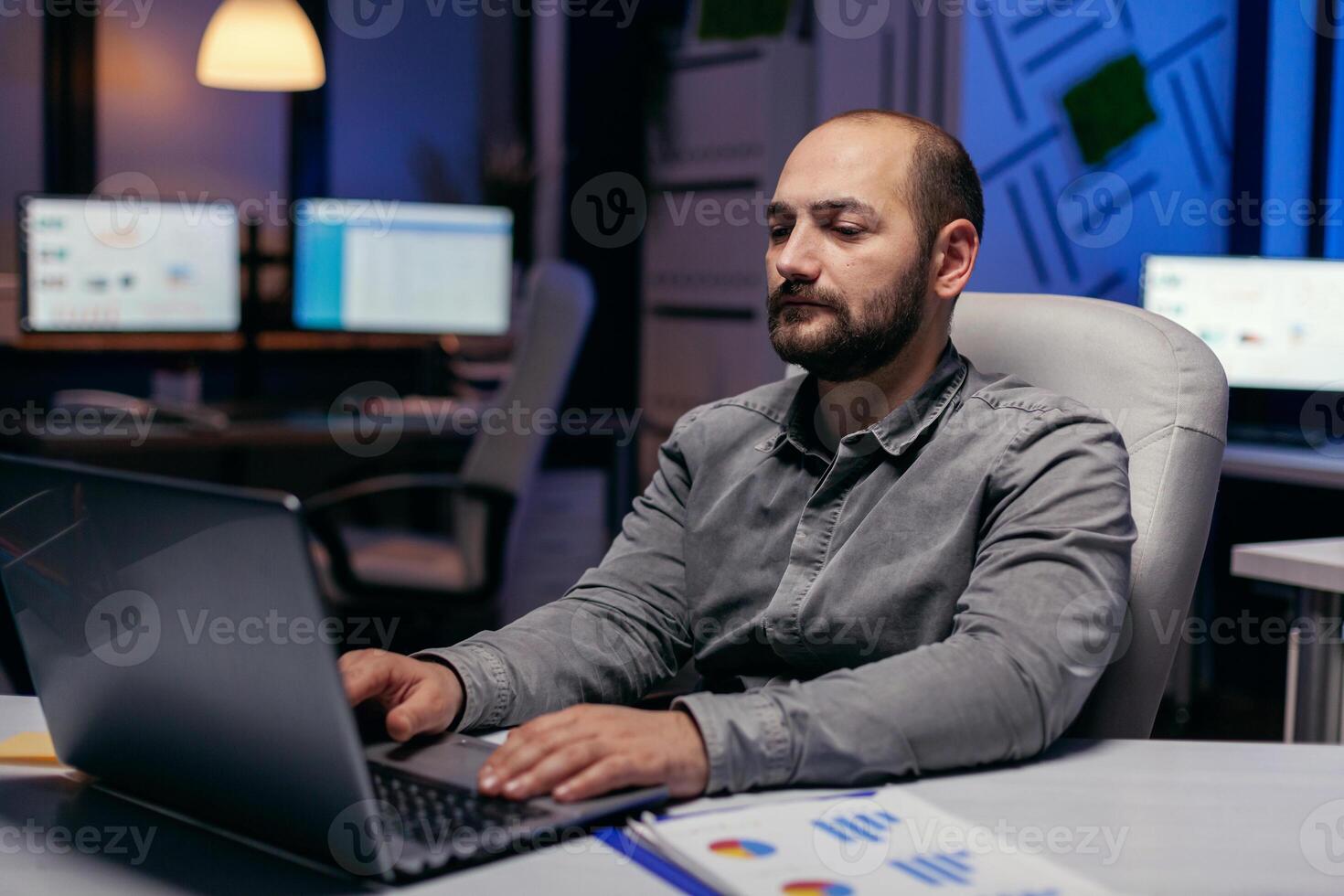 kaukasisch Unternehmer Arbeiten auf Frist im dunkel Büro Tippen auf Laptop. Clever Geschäftsmann Sitzung beim seine Arbeitsplatz im das Kurs von spät Nacht Std tun seine Arbeit. foto