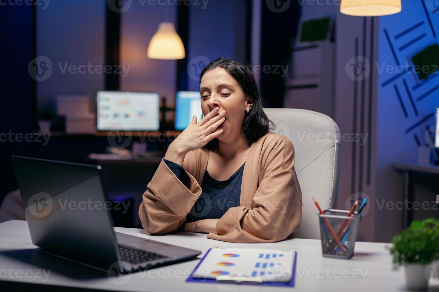 Unternehmer Dame gähnt Arbeiten auf Frist im dunkel Büro suchen beim Laptop Bildschirm. Clever Frau Sitzung beim ihr Arbeitsplatz im das Kurs von spät Nacht Std tun ihr Arbeit. foto