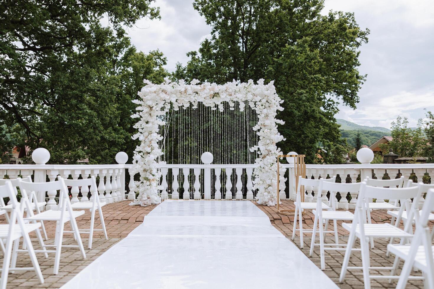 Hochzeit Bogen gemacht von Weiß Blumen auf ein Hintergrund von Bäume. ein Weiß Pfad zu das Bogen, viele Weiß Stühle. Vorbereitung zum das Hochzeit Zeremonie foto