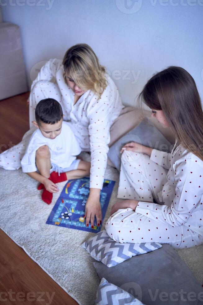 Mutter mit Teenager Tochter und wenig Sohn im Pyjama spielen Tafel Spiele auf das Fußboden foto