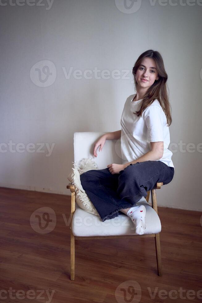 Porträt von ein schön Teenager Mädchen auf ein Stuhl im ein hell Zimmer im ein minimalistisch Stil foto