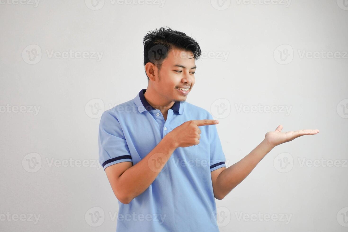 lächelnd asiatisch Mann mit ein öffnen Hand mit Finger zeigen zu das Seite tragen Blau t Hemd isoliert auf Weiß Hintergrund foto