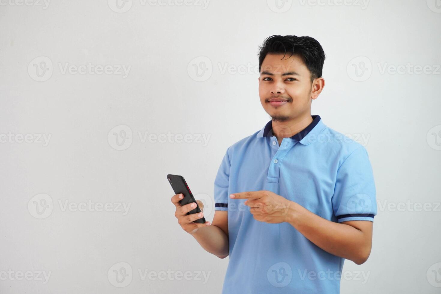 lächelnd oder glücklich jung asiatisch Mann tragen Blau t Hemd halten Telefon und zeigen beim Smartphone mit Finger, isoliert auf Weiß Hintergrund foto