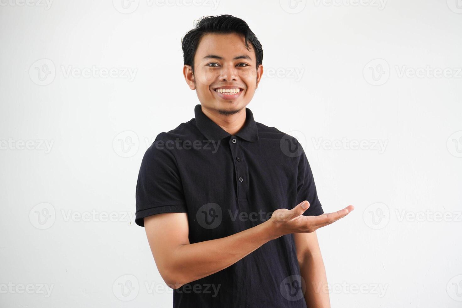 lächelnd jung asiatisch Mann im schwarz Polo Shirt, Weiß Hintergrund zeigen ein Kopieren Raum auf ein Palme und halten Ein weiterer Hand auf Taille. foto