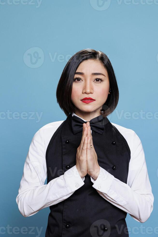 jung asiatisch Frau Rezeptionist meditieren mit gefaltet Palmen, entspannend und suchen beim Kamera. attraktiv Cafe Kellnerin tragen Uniform Stehen im Zen Pose Porträt auf Blau Hintergrund foto