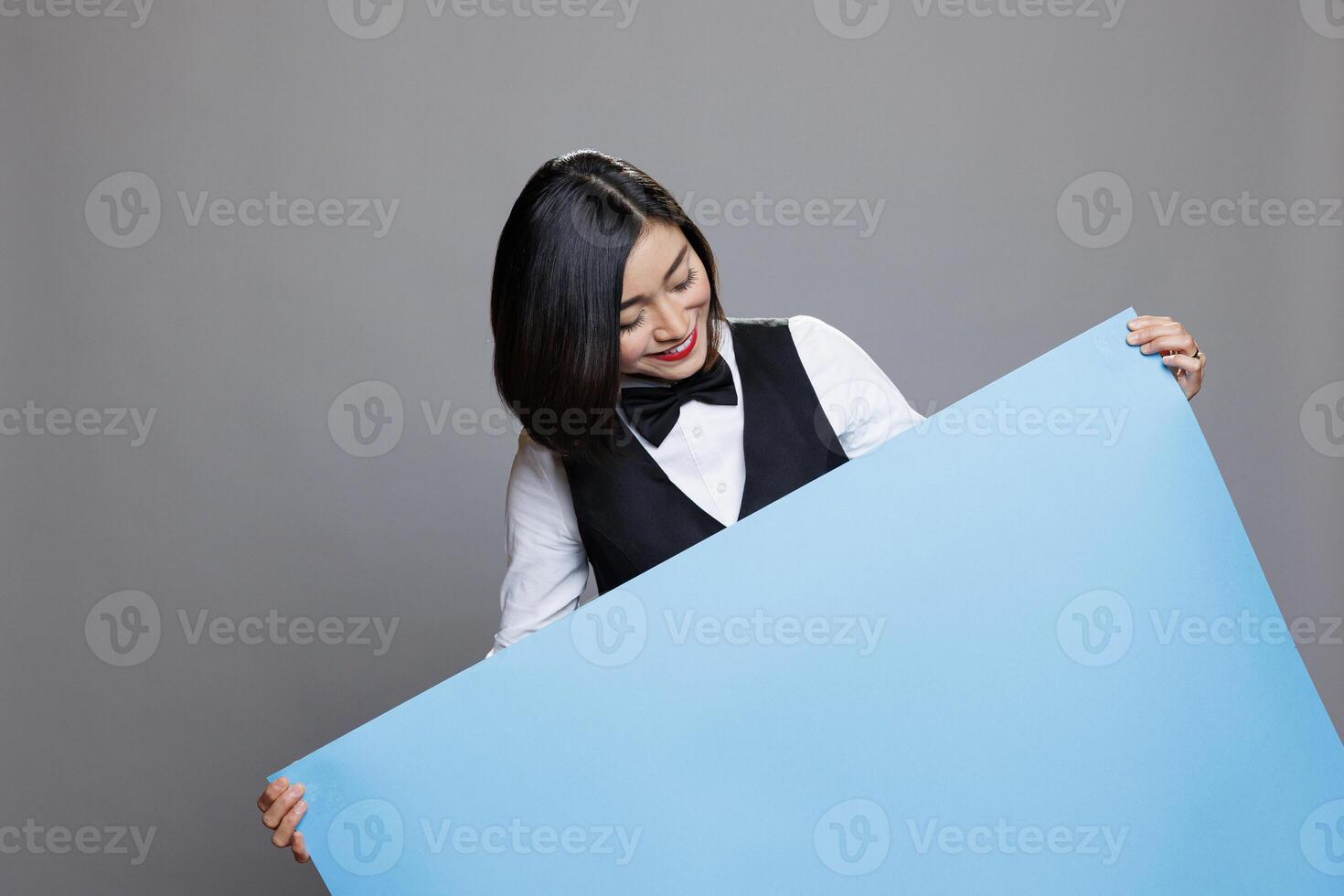 lächelnd asiatisch Kellnerin tragen Restaurant Uniform halten Blau leeren Papier Banner im Hände. Gastronomie Bedienung jung froh Frau Mitarbeiter posieren mit leer Beförderung Poster foto