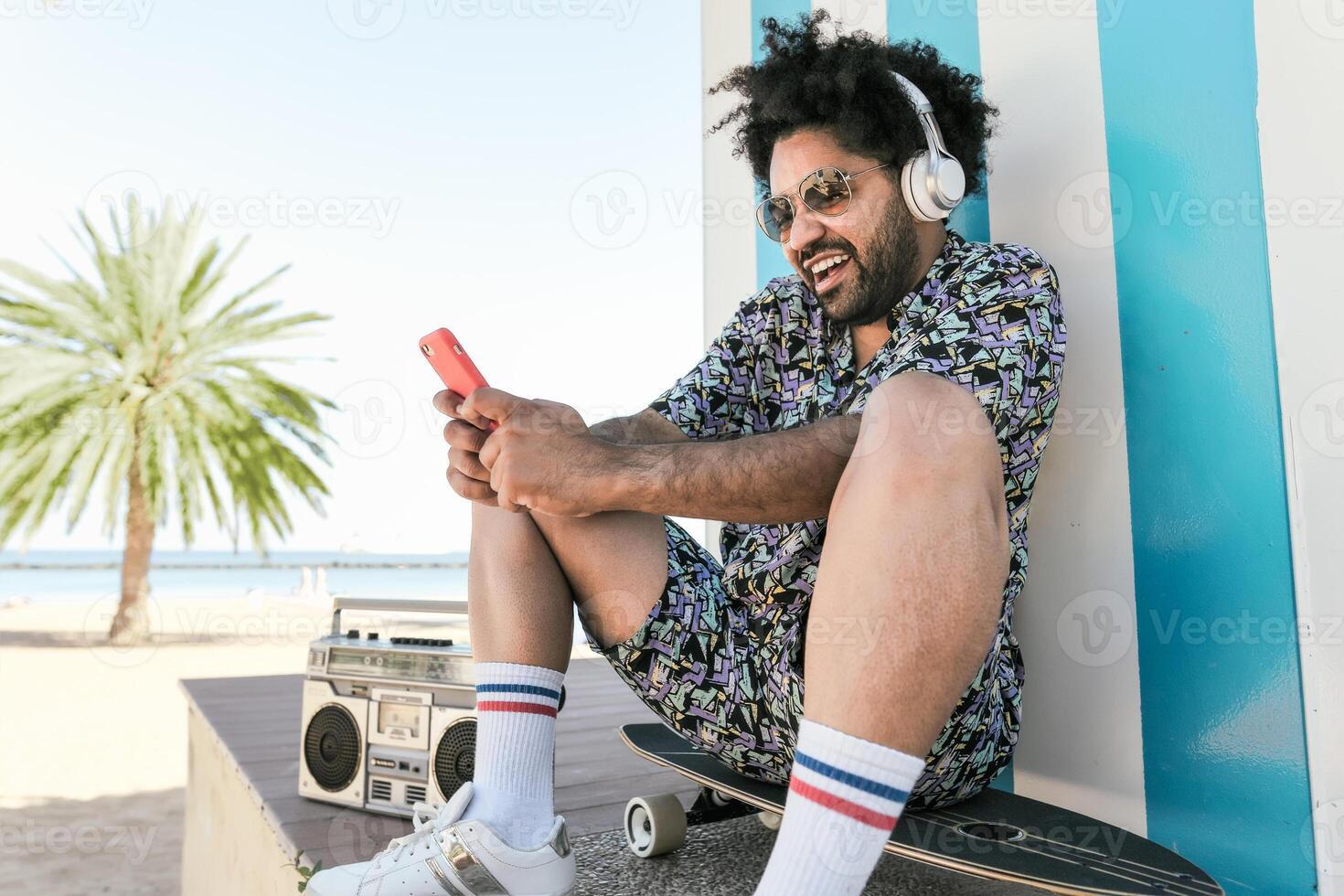 afro Latein Mann haben Spaß mit Handy, Mobiltelefon Smartphone und Hören Musik- mit Kopfhörer und Jahrgang Boombox Stereo auf tropisch Strand während Ferien Zeit foto