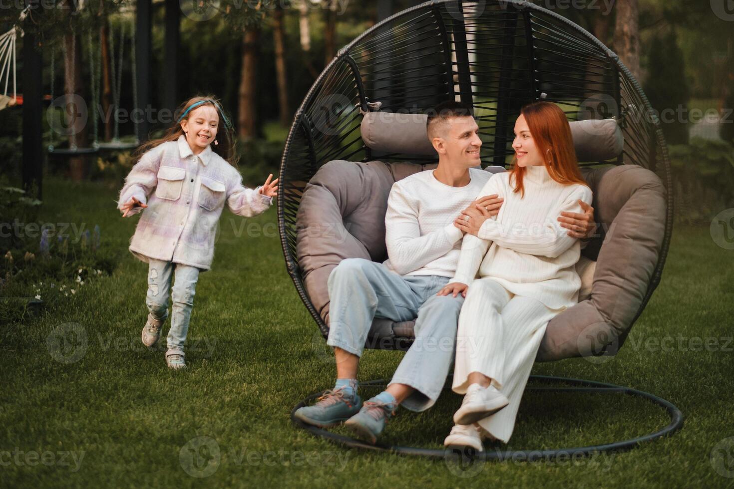 ein glücklich Familie ist Sitzung im ein Hängematte auf das Rasen in der Nähe von das Haus foto