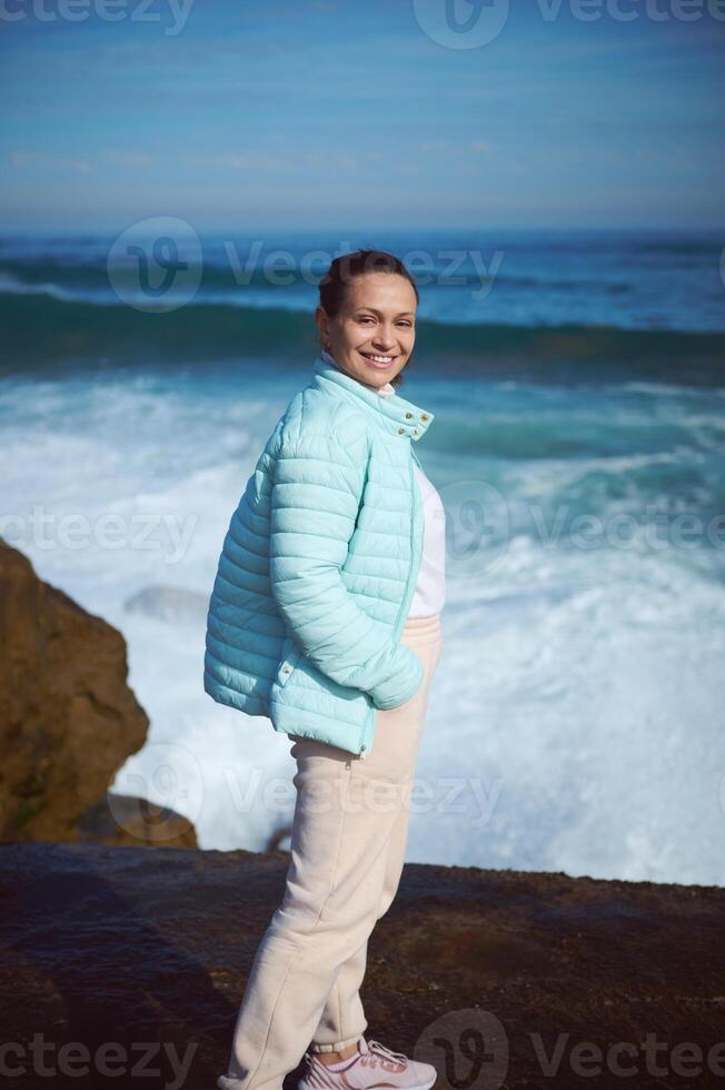 glücklich lächelnd Frau Stehen durch Ozean auf das felsig Cliff, suchen beim Kamera, genießen das Aussicht von schön Wellen foto