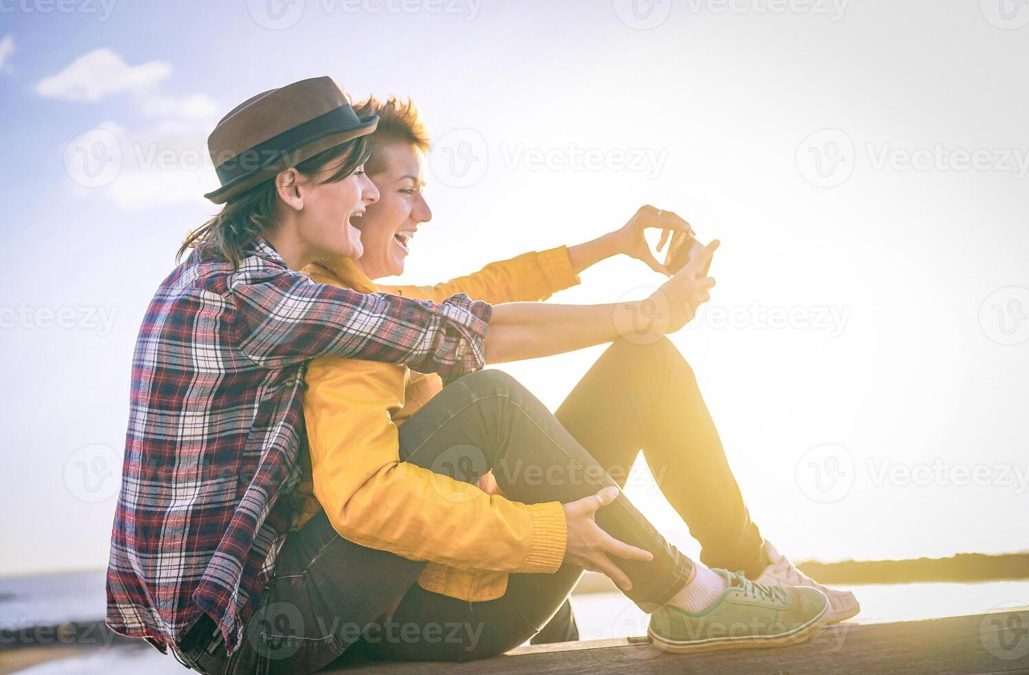 glücklich Lesben Paar nehmen ein Selfie mit Handy, Mobiltelefon Clever Telefon Kamera auf das Strand beim Sonnenuntergang - - Vignette bearbeiten - - Homosexualität, Diversität, Urlaub, reisen, LGBT, Technologie Konzept foto