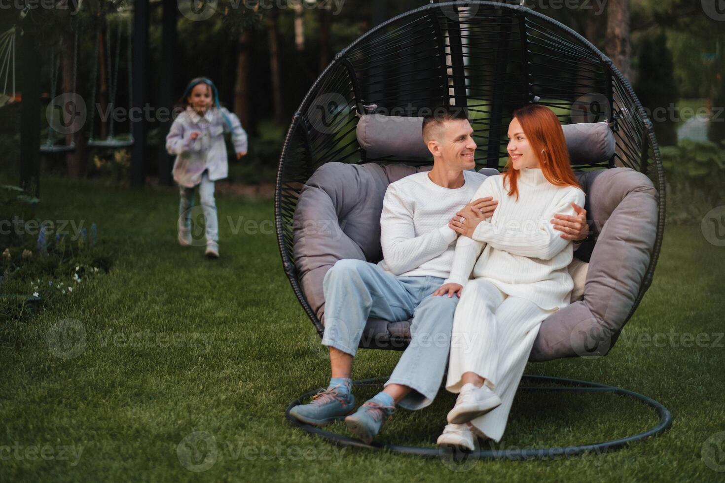 ein glücklich Familie ist Sitzung im ein Hängematte auf das Rasen in der Nähe von das Haus foto