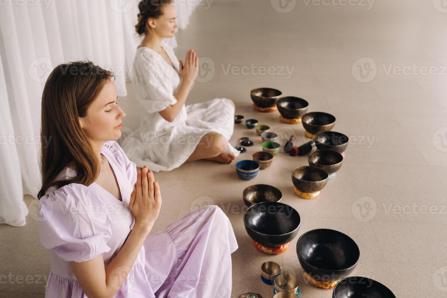 zwei Frauen sind Sitzung mit tibetanisch Schalen im das Lotus Position Vor ein Yoga Klasse im das Fitnessstudio foto