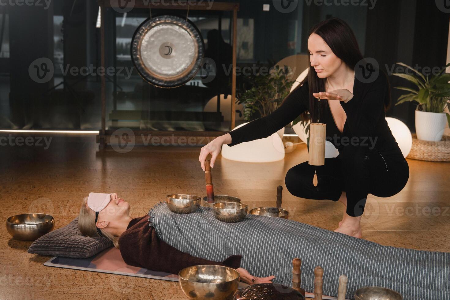 ein jung schön Frau ist tun ein Massage mit Singen Schalen und ein Koshi Glocke zu Ein weiterer Mädchen. Klang Therapie foto