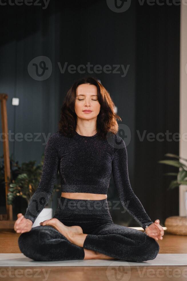 ein Frau im ein schwarz Trainingsanzug sitzt auf ein Yoga Matte im ein Lotus Position im das Fitnessstudio foto